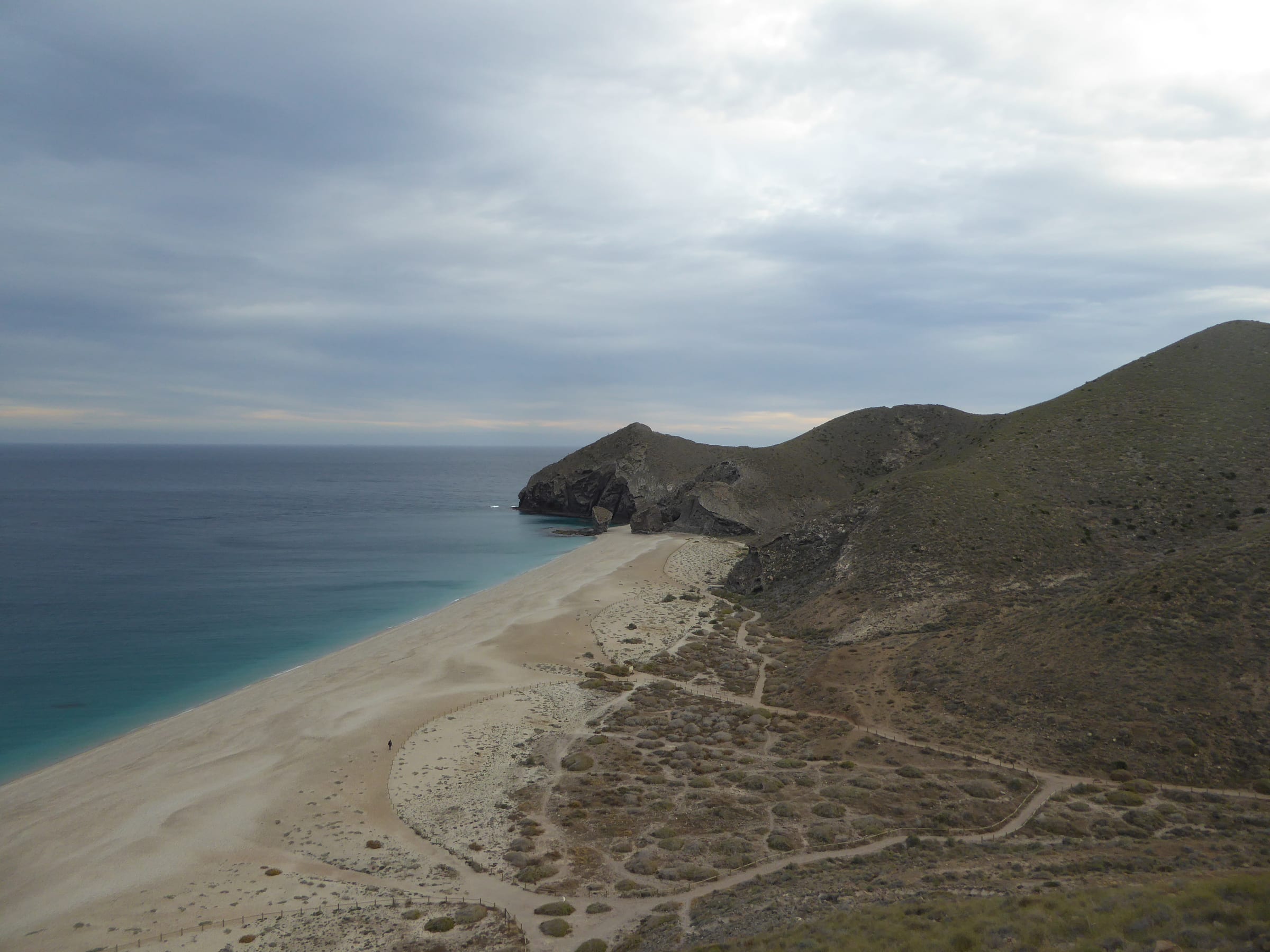 Long pale beach and clear blue-green water beneath rugged coastal hills at Playa de los Muertos, Spain.