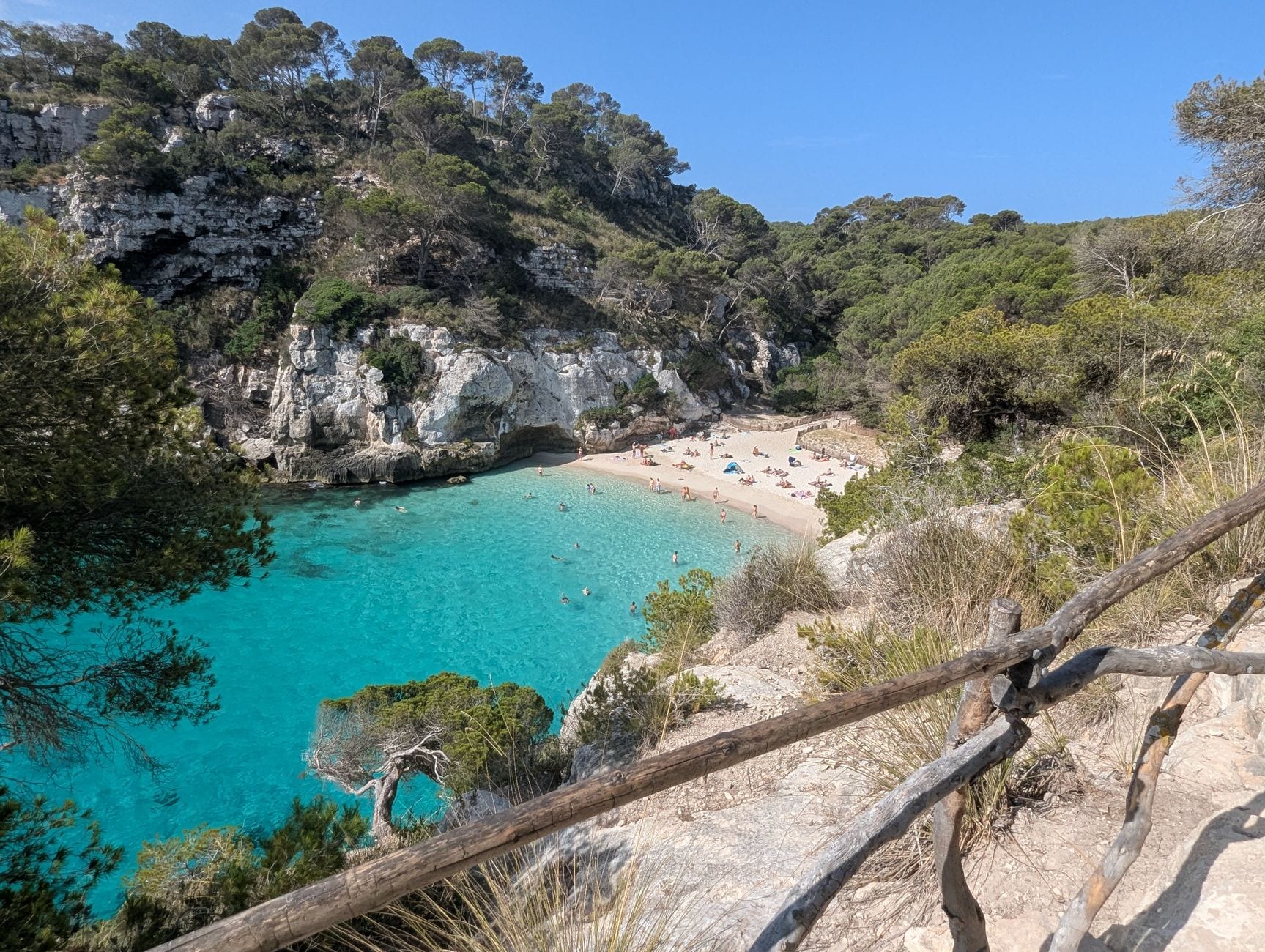 Cala Macarelleta’s white sand and turquoise water below a pine-covered clifftop path in Menorca