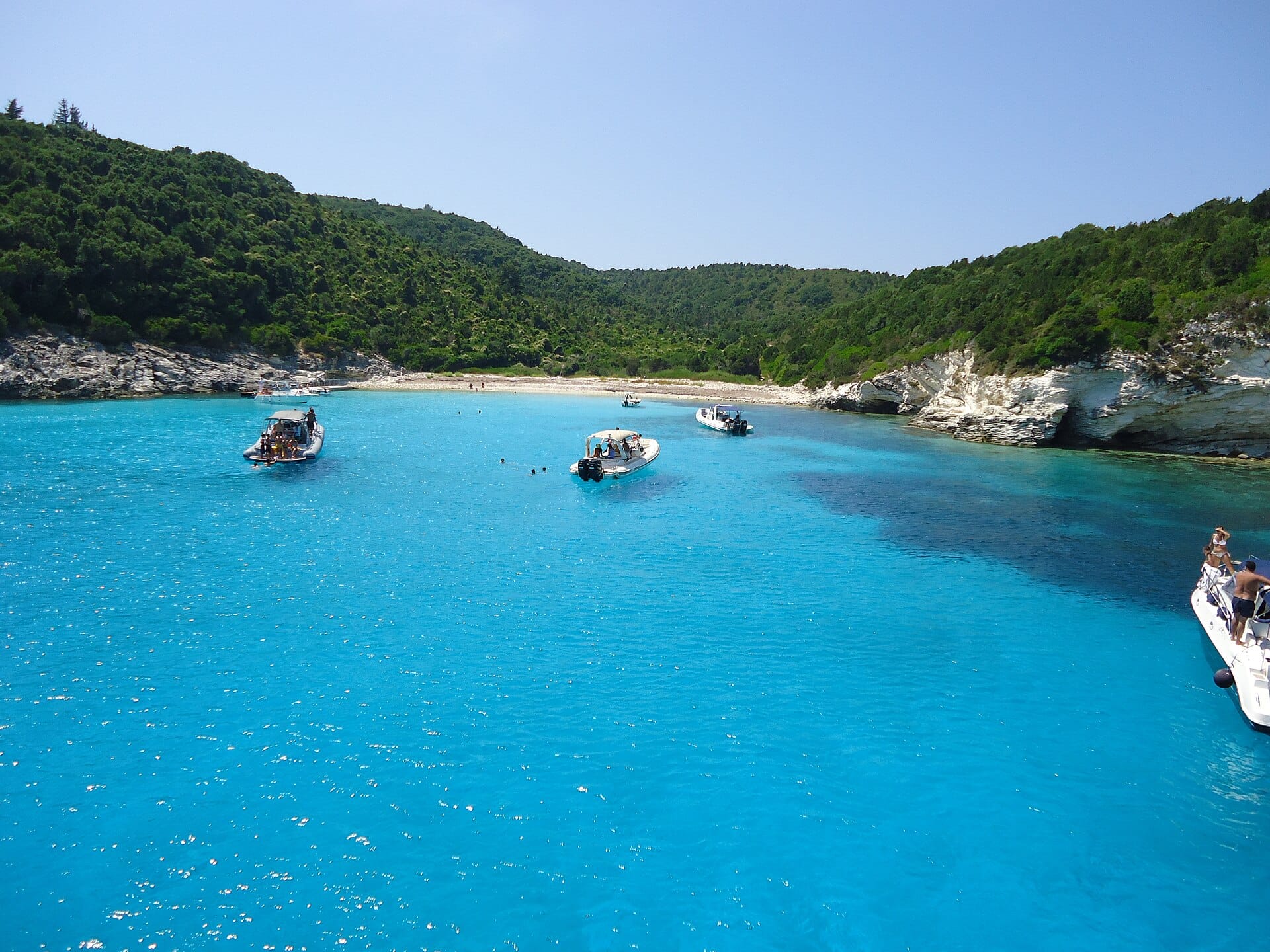 Motorboats anchored in bright turquoise water off Voutoumi beach, Antipaxos, below green hills
