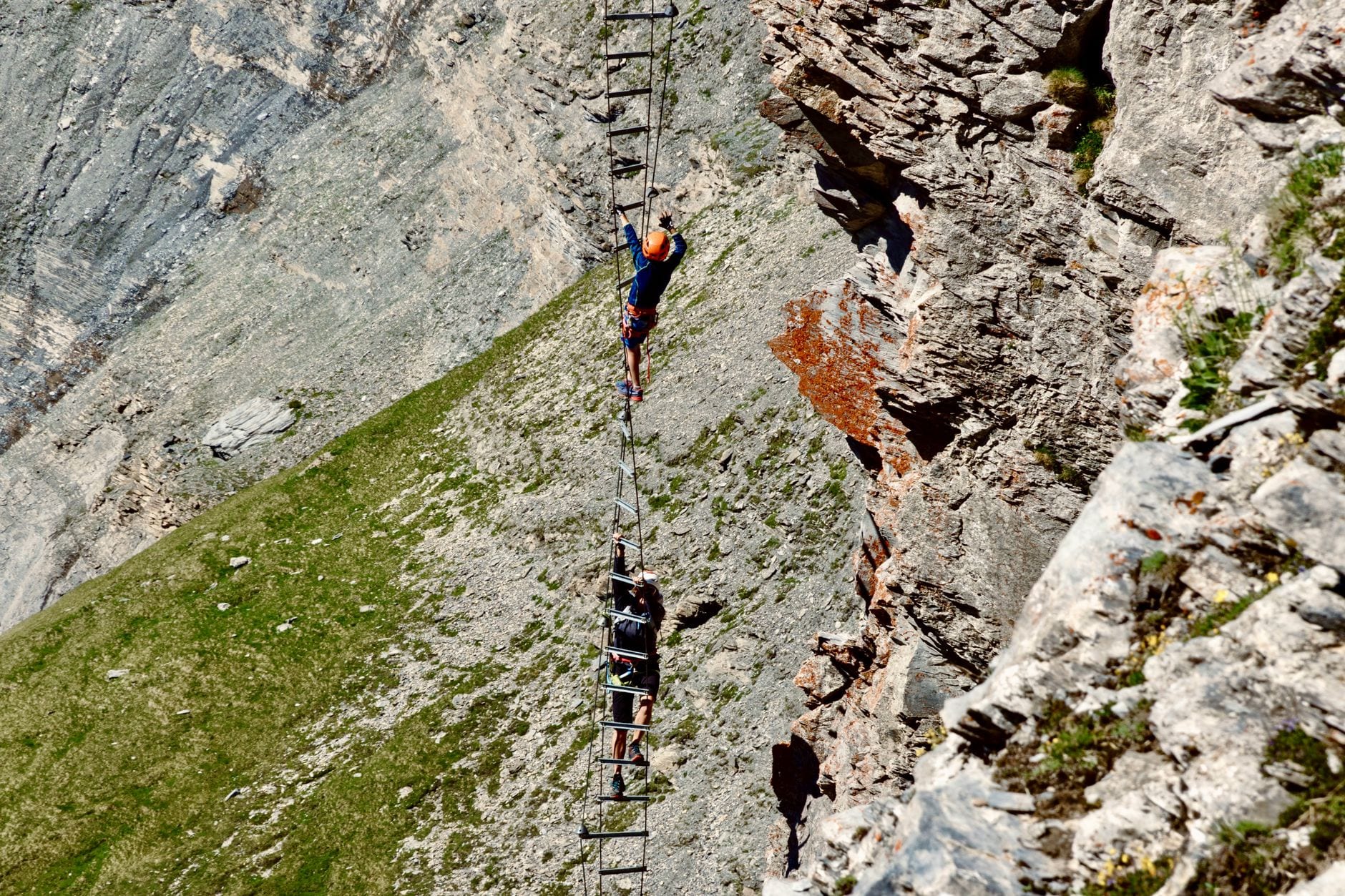 Two climbers in helmets and harnesses on a long metal ladder pitch of a via ferrata, above a steep grassy slope
